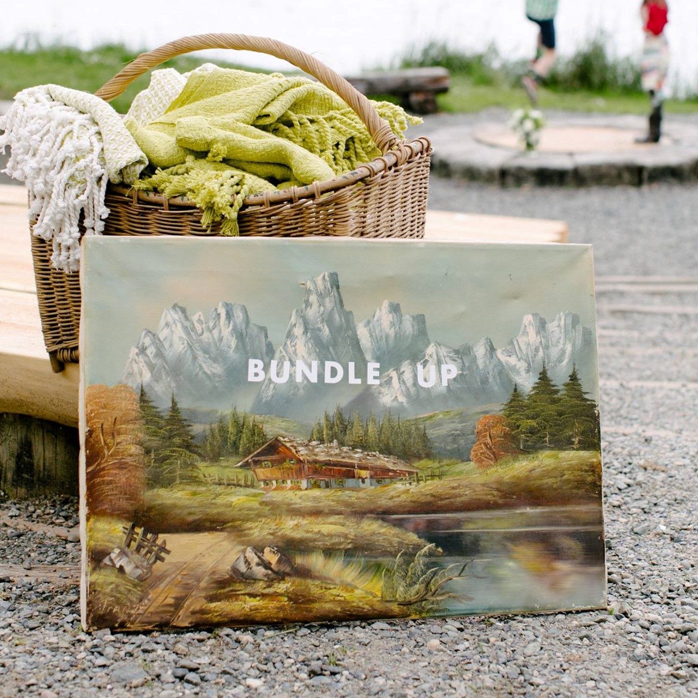 A basket of blankets behind a landscape painting with the words "Bundle up"